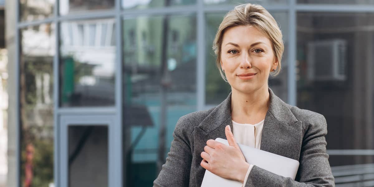 smiling professional woman in gray suit holding laptop in front of glass building